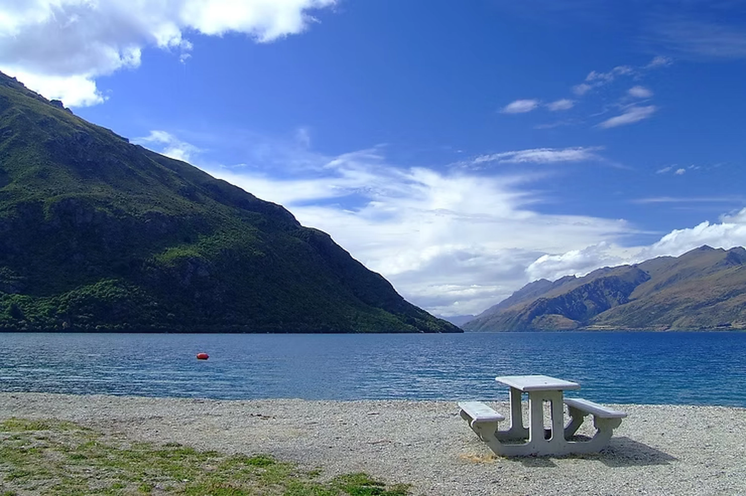 Wirtschaftsverein Apollon - Projektleitung von Konvaleszenz - Auf einem Kieselstrand am Rande eines großen, blauen Sees (wahrscheinlich Lake Wakatipu), umgeben von steilen, grünen Bergen unter einem strahlend blauen Himmel mit weißen Wolken, steht ein Picknicktisch aus Beton.