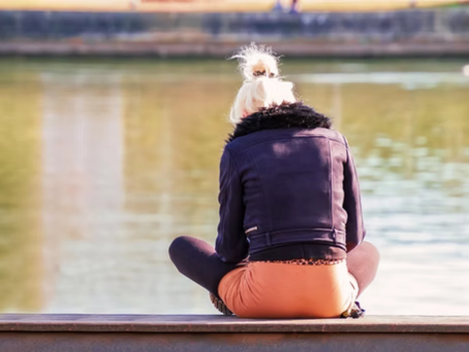 Wirtschaftsverein Apollon - Projektleitung von Konvaleszenz - Eine Person mit blonden, zu einem Dutt gebundenen Haaren, die eine dunkle Jacke und eine orangefarbene Hose trägt, sitzt im Schneidersitz auf einer Holzfläche und blickt auf ein Gewässer.