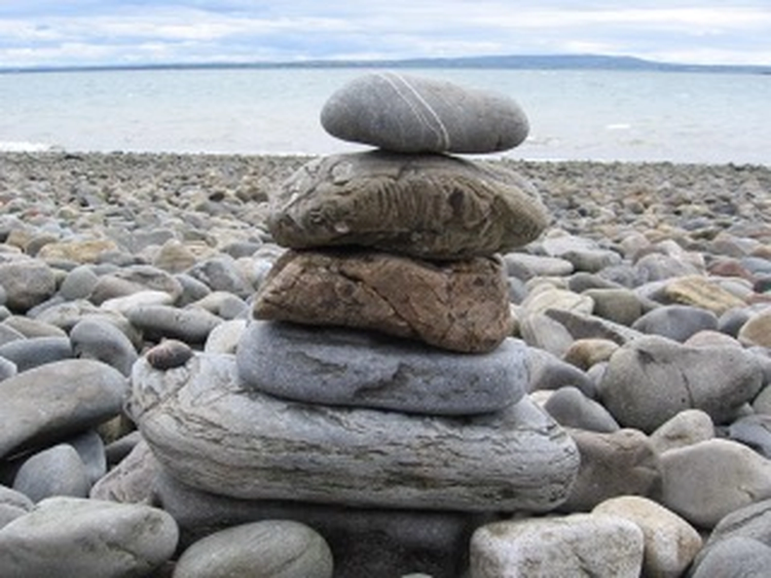 Wirtschaftsverein Apollon - Projektleitung von Konvaleszenz - Ein aus fünf glatten, grauen und braunen Steinen gestapelter Steinhaufen (ein Steinhaufen) balanciert auf einem Kieselstrand, im Hintergrund das Meer und der Horizont.